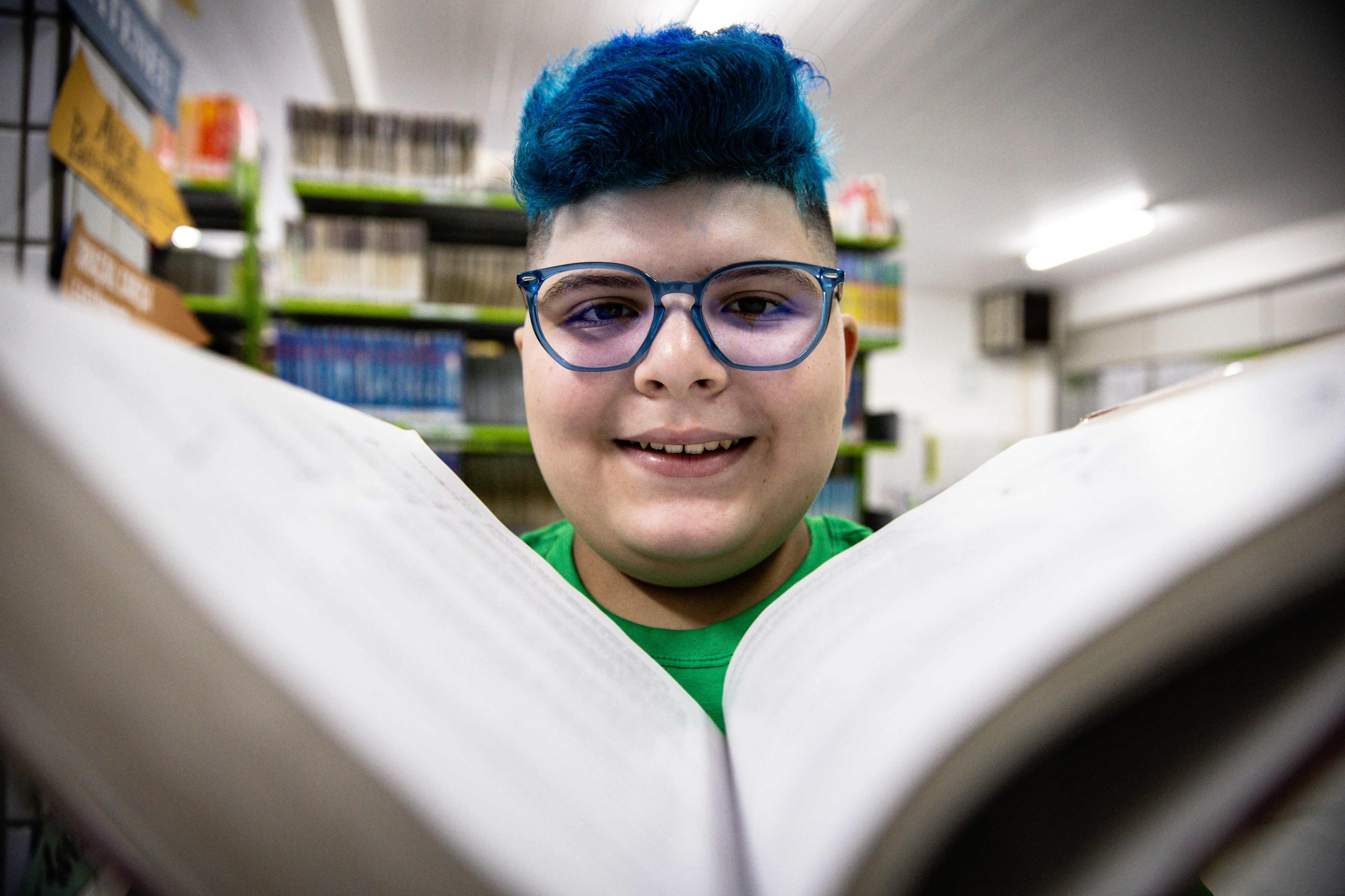 A imagem mostra um estudante sorridente, com cabelo azul vibrante e óculos azuis, a ler um livro aberto. A fotografia é tirada de baixo para cima, com o livro em primeiro plano, criando uma perspetiva que destaca o rosto do jovem. Ao fundo, estantes de livros sugerem que ele está numa biblioteca.