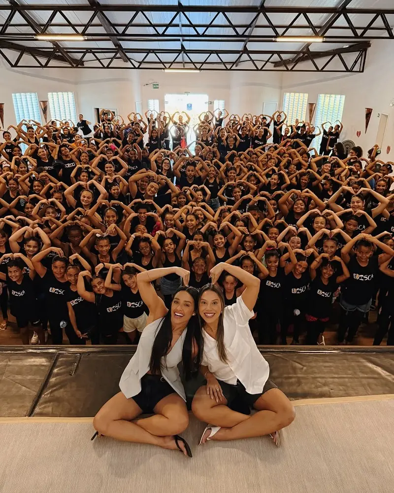 foto com várias crianças ao fundo e duas mulheres (Eva e Reanta do BBB 25) à frente fazendo um gestou sobre a cabeça