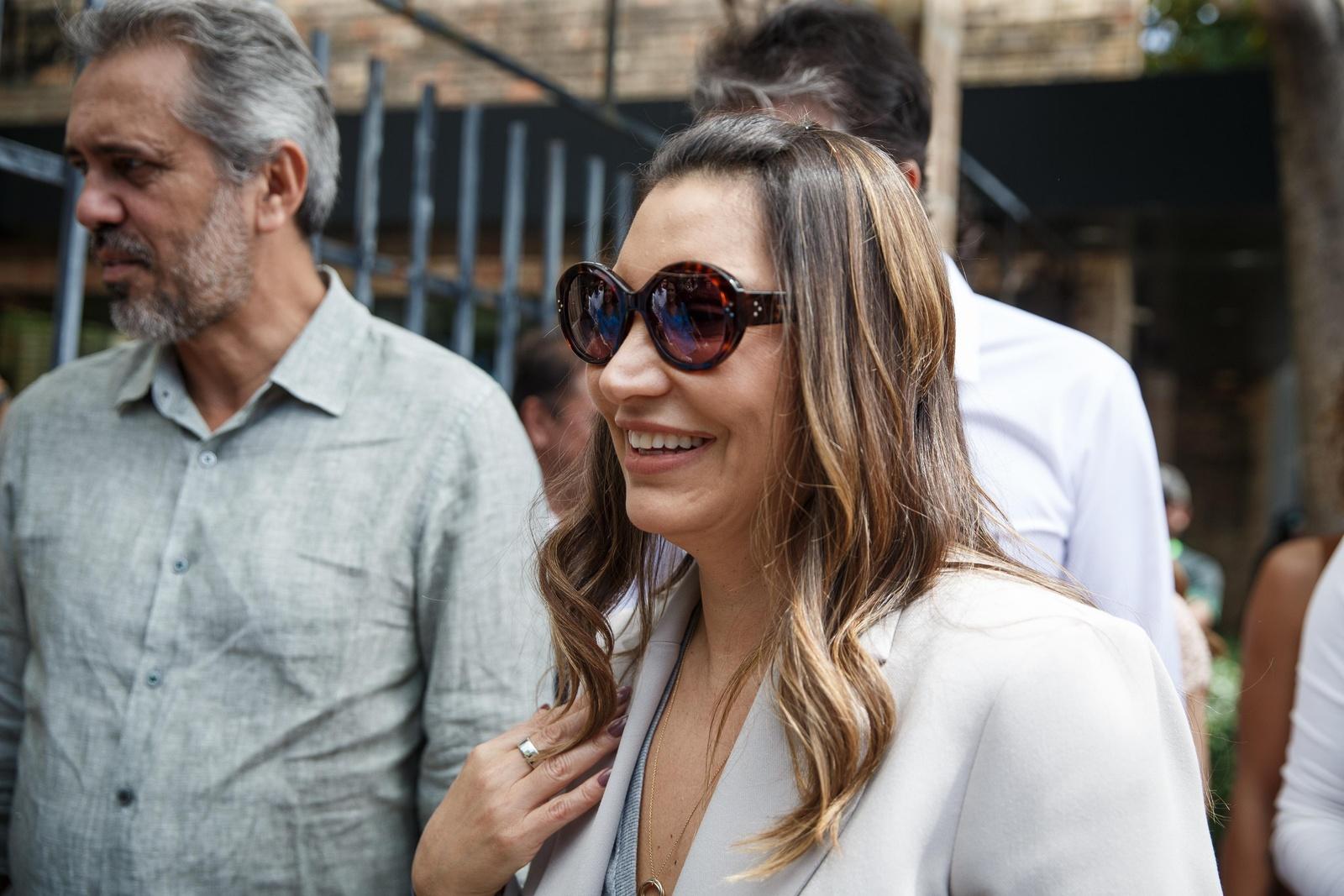 Imagem mostra a primeira-dama do Brasil, Janja Lula da Silva, vestida com um blazer cinza. Ela está sorrindo, tem cabelos castanhos com mechas loiras, de comprimento abaixo do ombro, e usa óculos escuros com armação marrom e lentes arredondadas. Ao fundo, aparece o governador do Ceará, Elmano de Freitas.