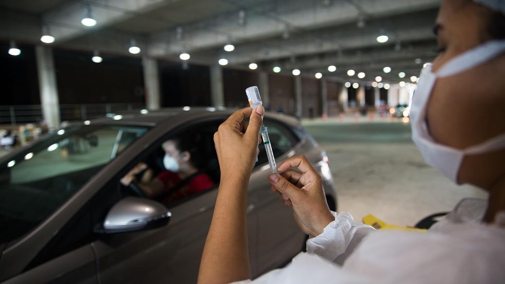 foto de drive-thru de vacinação em Fortaleza. A imagem ilustra uma enfermeira manuseando a vacina enquanto uma mulher aguarda no carro