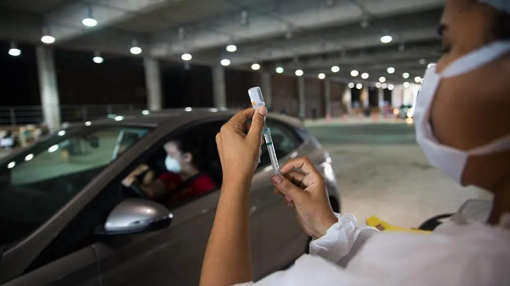 foto de drive-thru de vacinação em Fortaleza. A imagem ilustra uma enfermeira manuseando a vacina enquanto uma mulher aguarda no carro