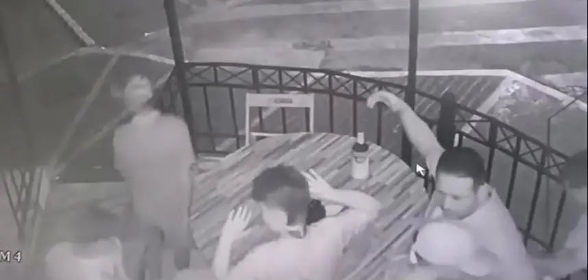 Imagem de uma cena noturna gravada por câmeras de segurança, mostrando três homens em uma mesa de pôquer ao ar livre, com uma cerca de ferro ao fundo. A imagem está em preto e branco.