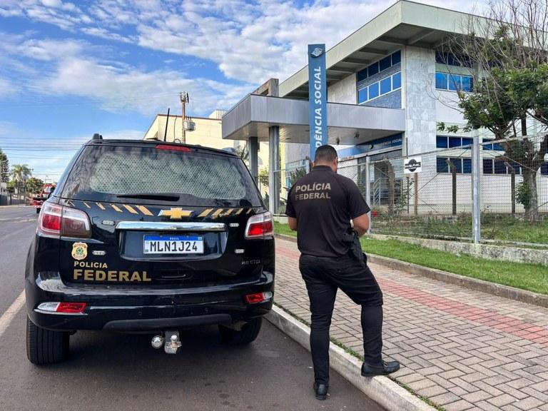 Polícia Federal na frente de uma unidade de previdência social, com um carro oficial parado na rua, e um policial de costas olhando para a entrada do prédio.