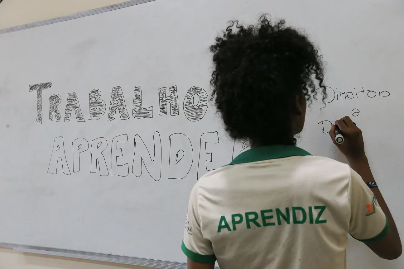 Foto de menina negra de cabelos cacheados, de costas, com blusa de aprendiz, escrevendo a frase Trabalho e Aprender num quadro branco