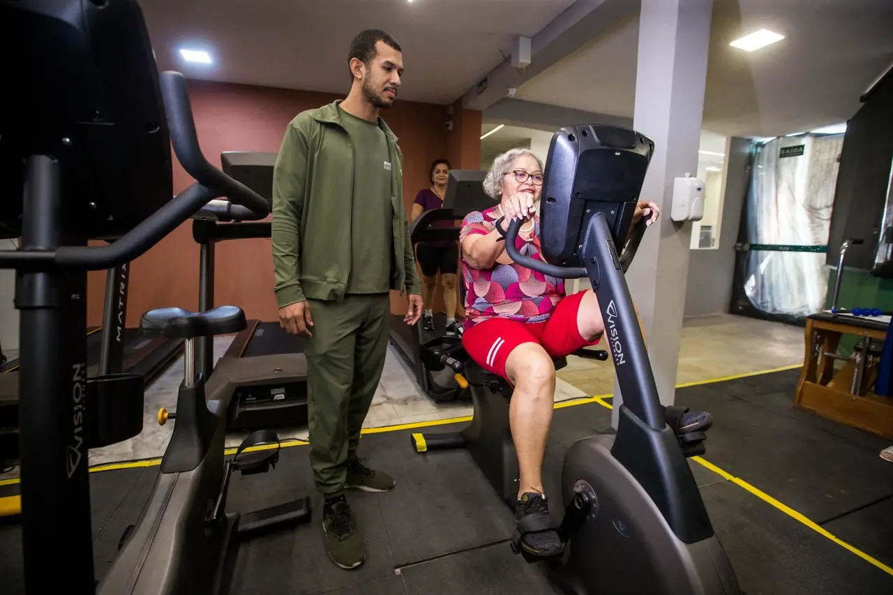 Foto mostra idosa usando aparelho de musculação enquanto é acompanhada por um profissional de Educação Física para matéria sobre apps de bem-estar