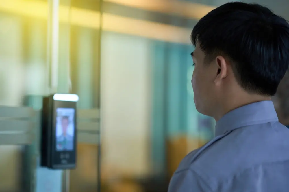 A imagem mostra um homem de perfil, de frente para um dispositivo de segurança com reconhecimento facial instalado em uma porta de vidro. O rosto do homem está parcialmente visível e seu olhar está direcionado para o dispositivo.