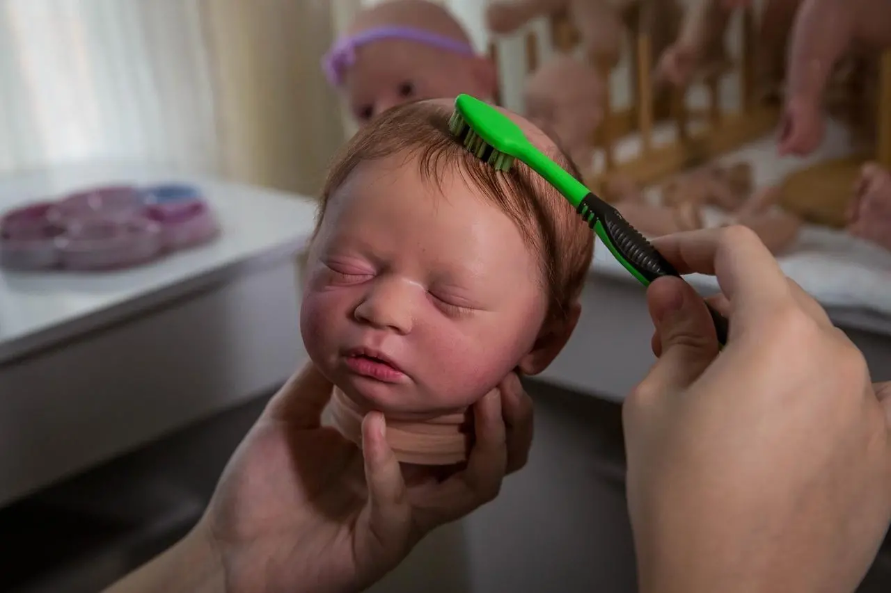 Imagem mostra a cabeça de um bebê reborn, boneco que simula um bebê real. O boneco está de olhos fechados e tem cabelos finos sendo penteados com uma escova de dentes verde e preta.