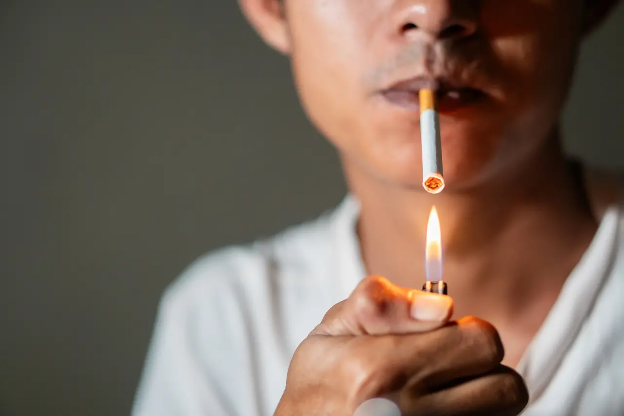 Imagem mostra homem jovem acendendo e fumando cigarro tradicional, sobre fundo escuro, ilustrando como parar de fumar em dia mundial sem tabaco