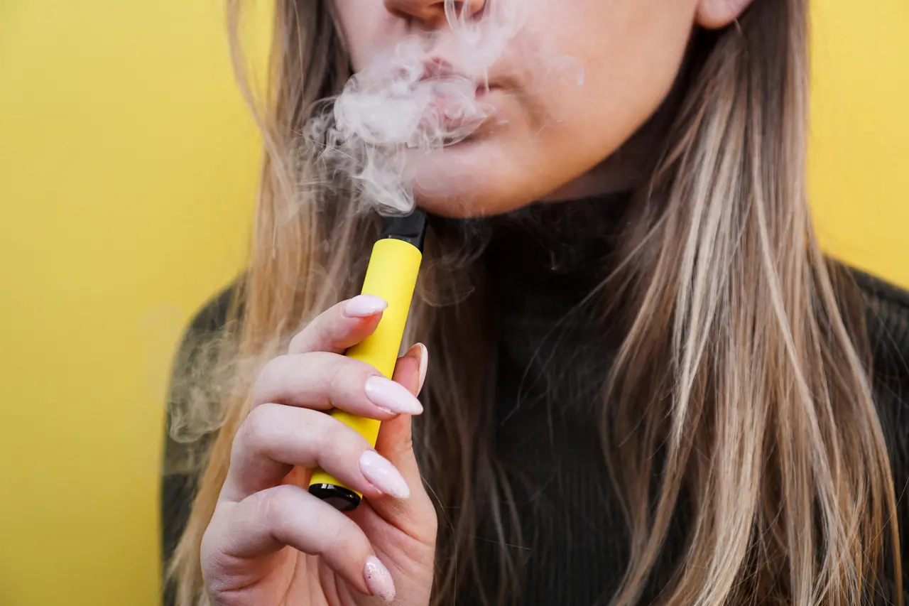 Imagem mostra mulher jovem fumando cigarro eletrônico (ou vape, ou pod), sobre fundo amarelo, ilustrando como parar de fumar em dia mundial sem tabaco