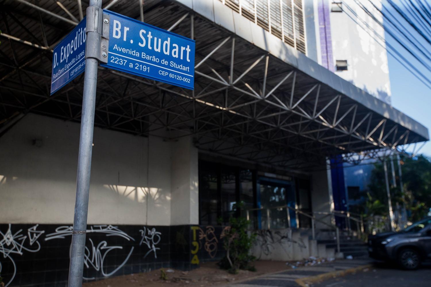Foto que contém placa do cruzamento da avenida Barão de Studart com a rua Dom Expedito Lopes, em Fortaleza. Ao fundo, prédio abandonado da Caixa Econômica Federal