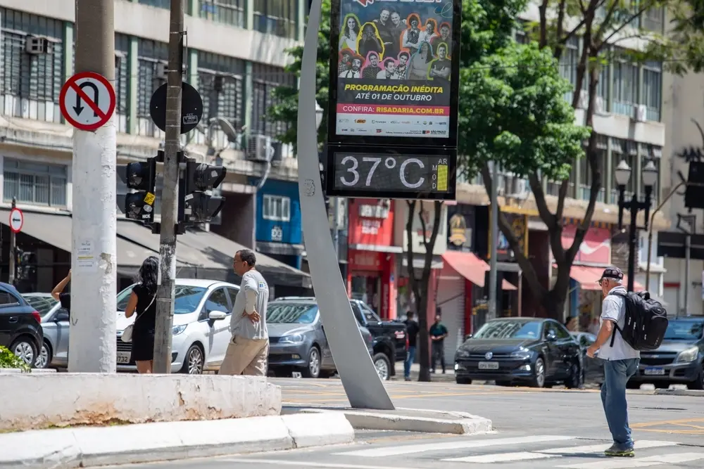 Imagem de uma rua movimentada em uma cidade com um painel eletrônico exibindo a temperatura de 37°C, com pedestres esperando para atravessar a rua, árvores e edifícios ao fundo.