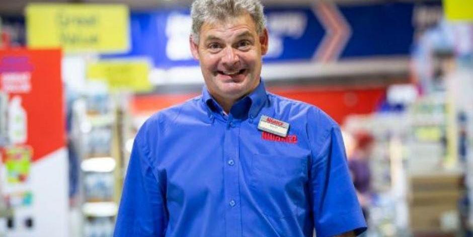 Funcionário sorridente de loja de varejo em um supermercado, vestindo uniforme azul, promovendo atendimento ao cliente e confiabilidade no setor de varejo.