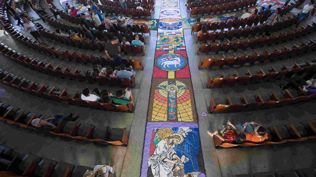 Vista aérea do interior de uma igreja com bancos dispostos em semicírculo e um tapete colorido feito com serragem no corredor central. O tapete apresenta imagens religiosas, como Jesus crucificado, símbolos cristãos e cenas bíblicas, em comemoração ao Corpus Christi. Fieis estão sentados nos bancos, observando o tapete artístico.
