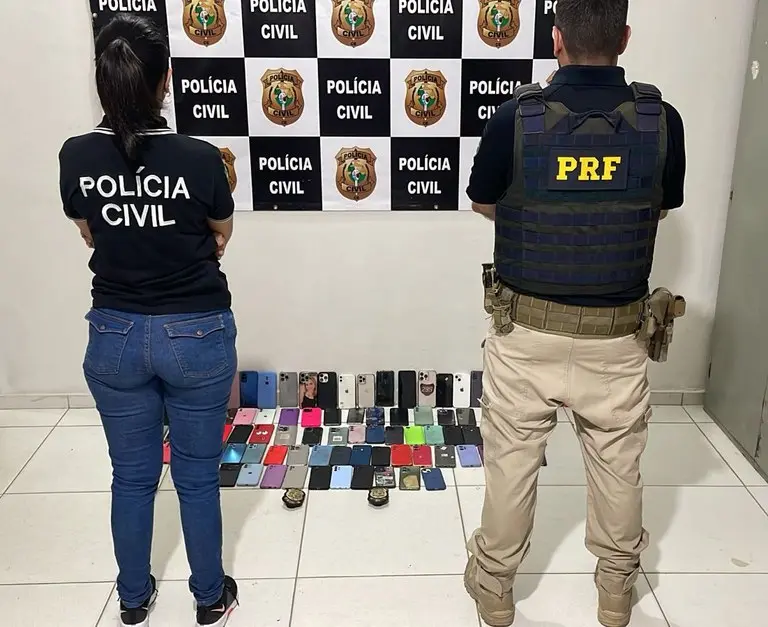 Imagem de dois agentes policiais, um da Polícia Civil e outro da Polícia Rodoviária Federal, com diversos celulares recuperados