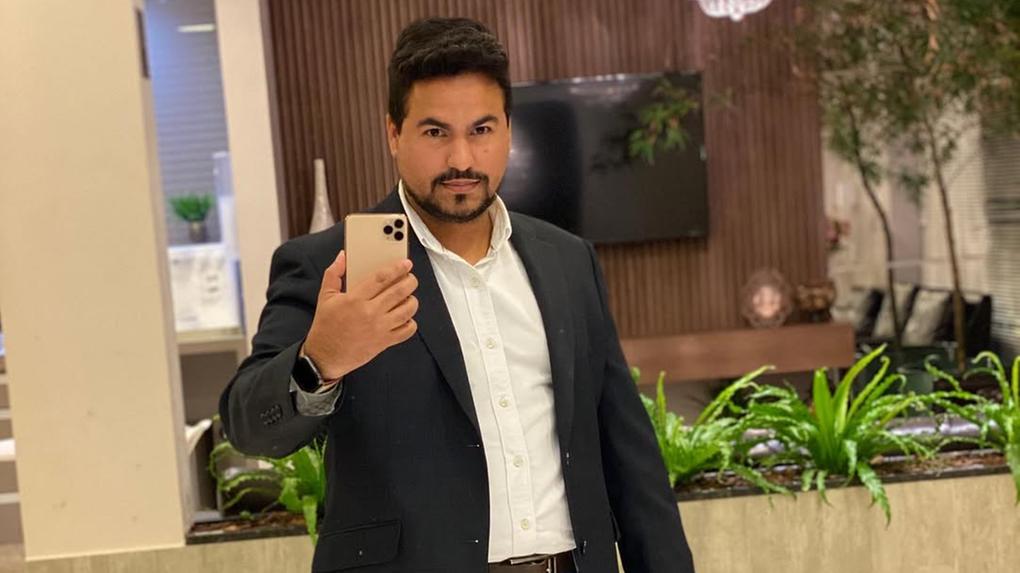 Homem de terno e jeans tirando uma selfie em ambiente elegante com decoração moderna e plantas verdes ao fundo.