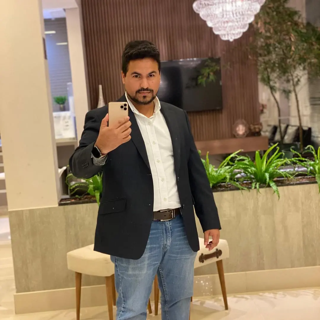 Homem de terno e jeans tirando uma selfie em ambiente elegante com decoração moderna e plantas verdes ao fundo.