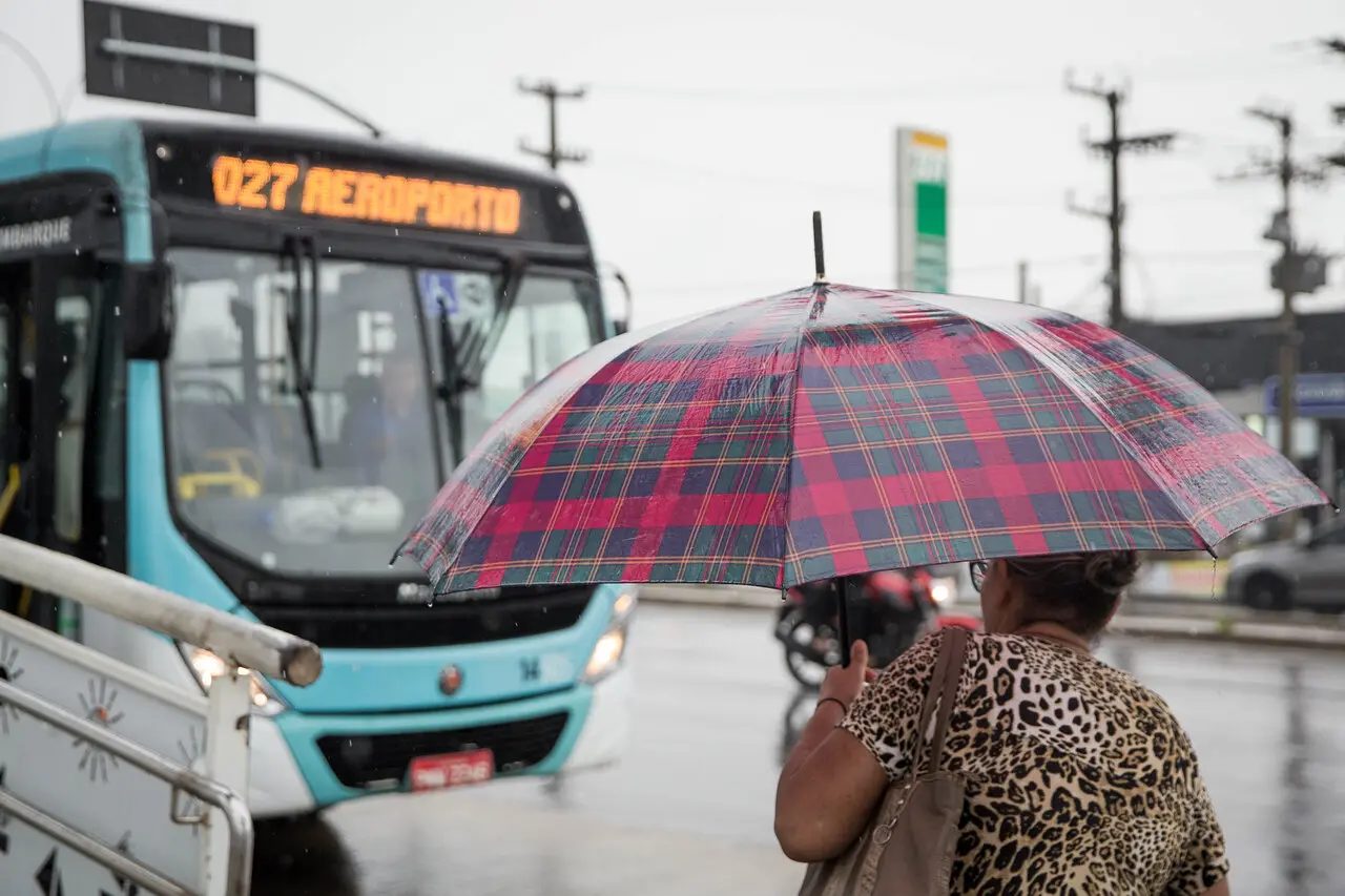 Pessoa com guarda-chuva em dia de chuva na rua, próxima a um ônibus urbano em Fortaleza, onde amanheceu chovendo nesta terça-feira (3), apesar do fim da quadra chuvosa.