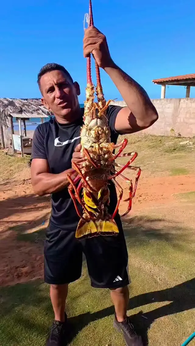 Homem segurando uma lagosta gigante ao ar livre com céu azul ao fundo, em Icapuí, no Ceará, próximo à praia ou área costeira.
