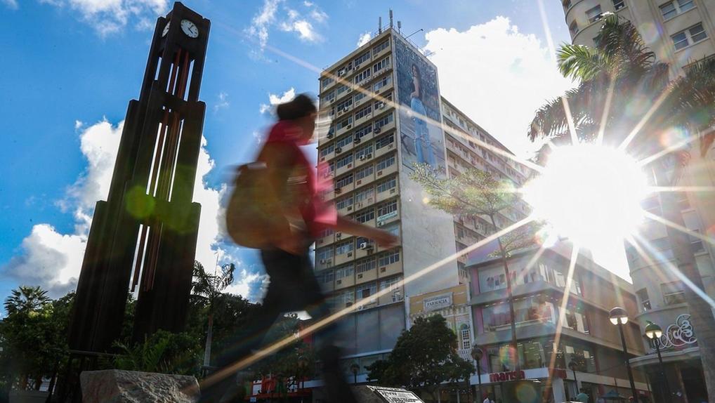 A imagem retrata um dia ensolarado na Praça do Ferreira, em Fortaleza. Em primeiro plano, uma pessoa em movimento está embaçada, vestindo calça escura, camisa rosa e carregando uma mochila, atravessando um chão de pedras irregulares. À esquerda, uma alta torre de relógio escura se destaca contra o céu azul com nuvens brancas. Ao fundo, vários edifícios altos com janelas visíveis compõem o cenário urbano. O sol brilha intensamente no lado direito da imagem, criando um forte brilho e raios de luz visíveis. Palmeiras e outra vegetação complementam a cena.