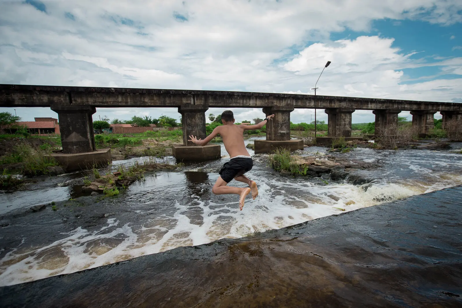 Imagem captura o momento em que um jovem, sem camisa e de calção escuro, salta no ar sobre um rio com águas agitadas e espumosas. Seus braços estão abertos e as pernas dobradas, indicando o movimento do salto. Ao fundo, uma ponte antiga e robusta, com pilares de concreto, atravessa o açude São Pedro Timbaúba, no interior do Ceará. As margens são cobertas por vegetação e, à distância, algumas construções baixas podem ser vistas. O céu é parcialmente nublado, com nuvens brancas espalhadas.