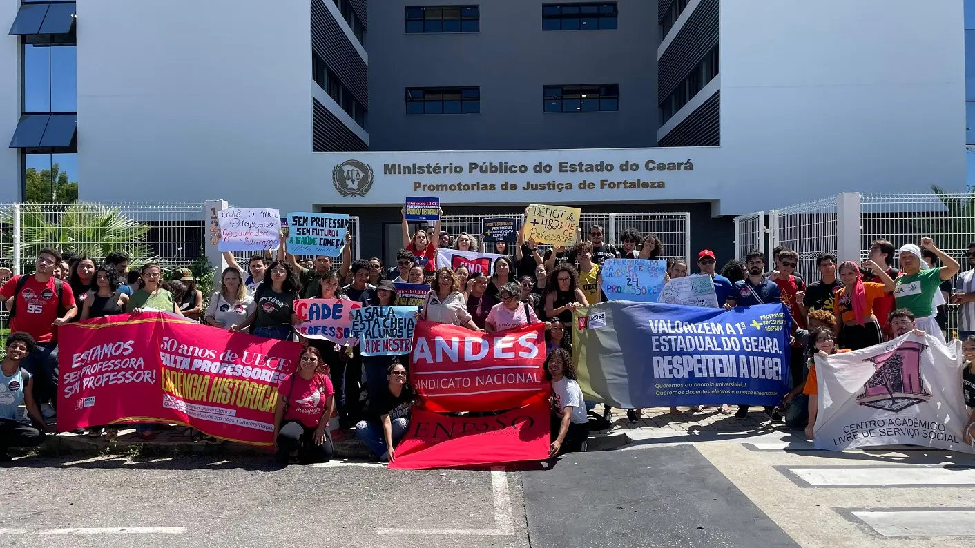 Foto de um protesto em frente ao Ministério Público do Estado do Ceará, em Fortaleza. Um grupo de estudantes e professores segura faixas e cartazes coloridos exigindo mais professores, investimentos e respeito à Universidade Estadual do Ceará (UECE). As mensagens destacam a carência histórica de docentes e a valorização da educação pública estadual.