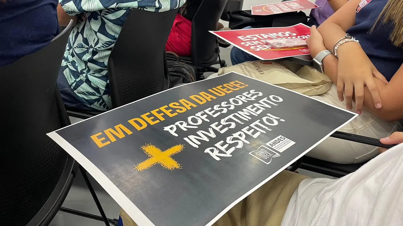 Foto de cartaz preto com o nome Em defesa da Uece + professores, investimentos e respeito sobre uma carteira em uma sala fechada