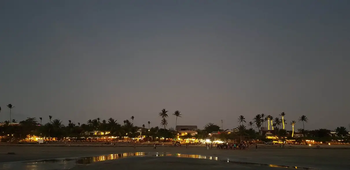 Foto da praia de Jericoacoara à noite, com luzes amarelas e quentes iluminando bares e restaurantes à beira-mar. Silhuetas de coqueiros contrastam com o céu escuro, e pessoas aproveitam o ambiente noturno na areia.