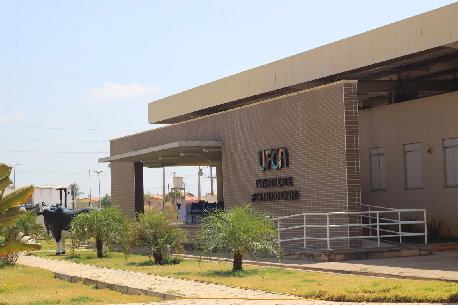 Imagem mostra a fachada da Universidade Federal do Cariri, no Ceará. Em primeiro plano, aparece um pequeno jardim com plantas. Atrás, o letreiro da UFCA. A fachada é em cores marrom e bege.
