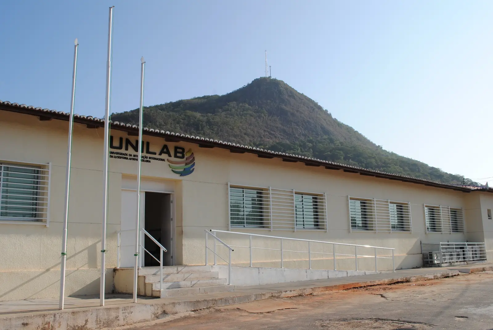 Imagem mostra a fachada do campus da Unilab em Redenção, no Ceará, amarela com janelas de grades brancas. Atrás aparece uma serra coberta por vegetação verde.