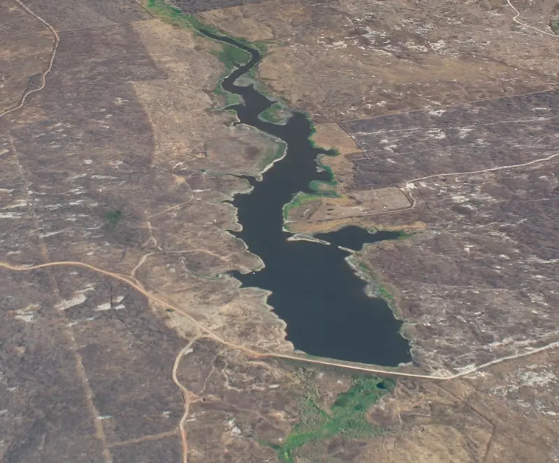 A imagem é uma fotografia aérea vertical que mostra um reservatório de água cercado por uma paisagem predominantemente árida e seca. Um corpo d'água escuro, em tons de azul e verde-escuro, que se estende de forma sinuosa na vertical, ocupando a maior parte do centro da imagem. O reservatório é ladeado por uma paisagem seca, de coloração predominantemente marrom-claro e bege.