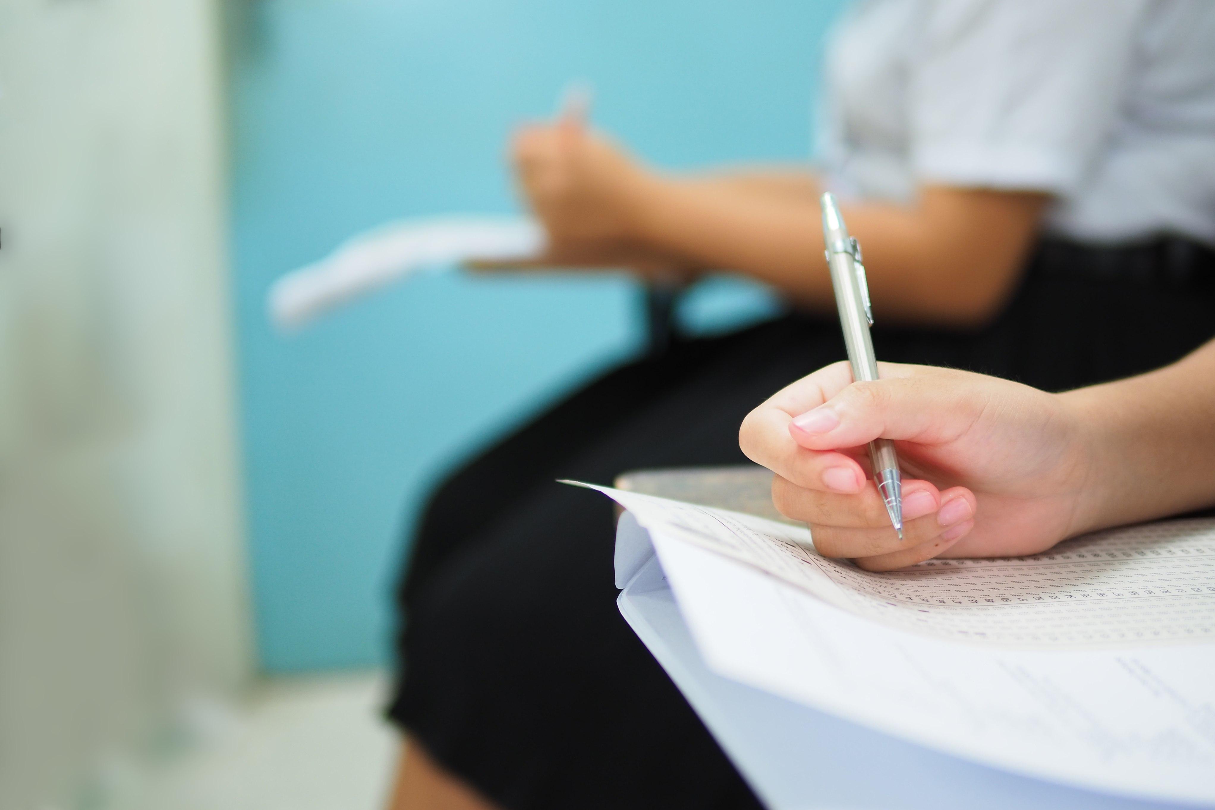 Foto de uma pessoa segurando uma caneta e fazendo uma prova em uma sala de aula