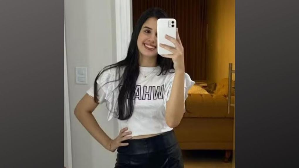 Karen Marques Abreu sorridente fazendo selfie em espelho com telefone celular, usando camiseta branca e saia preta, em ambiente de quarto com decoração moderna.