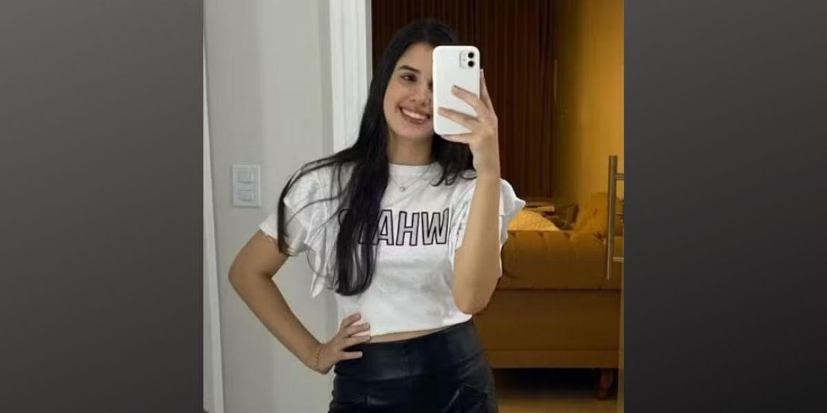 Karen Marques Abreu sorridente fazendo selfie em espelho com telefone celular, usando camiseta branca e saia preta, em ambiente de quarto com decoração moderna.