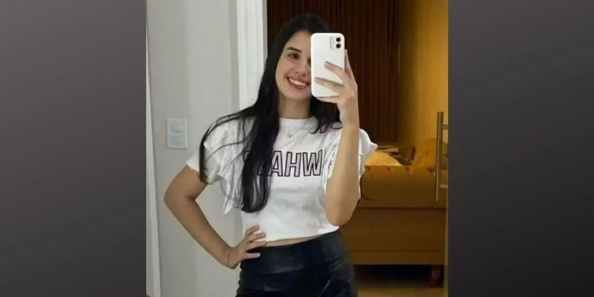 Karen Marques Abreu sorridente fazendo selfie em espelho com telefone celular, usando camiseta branca e saia preta, em ambiente de quarto com decoração moderna.