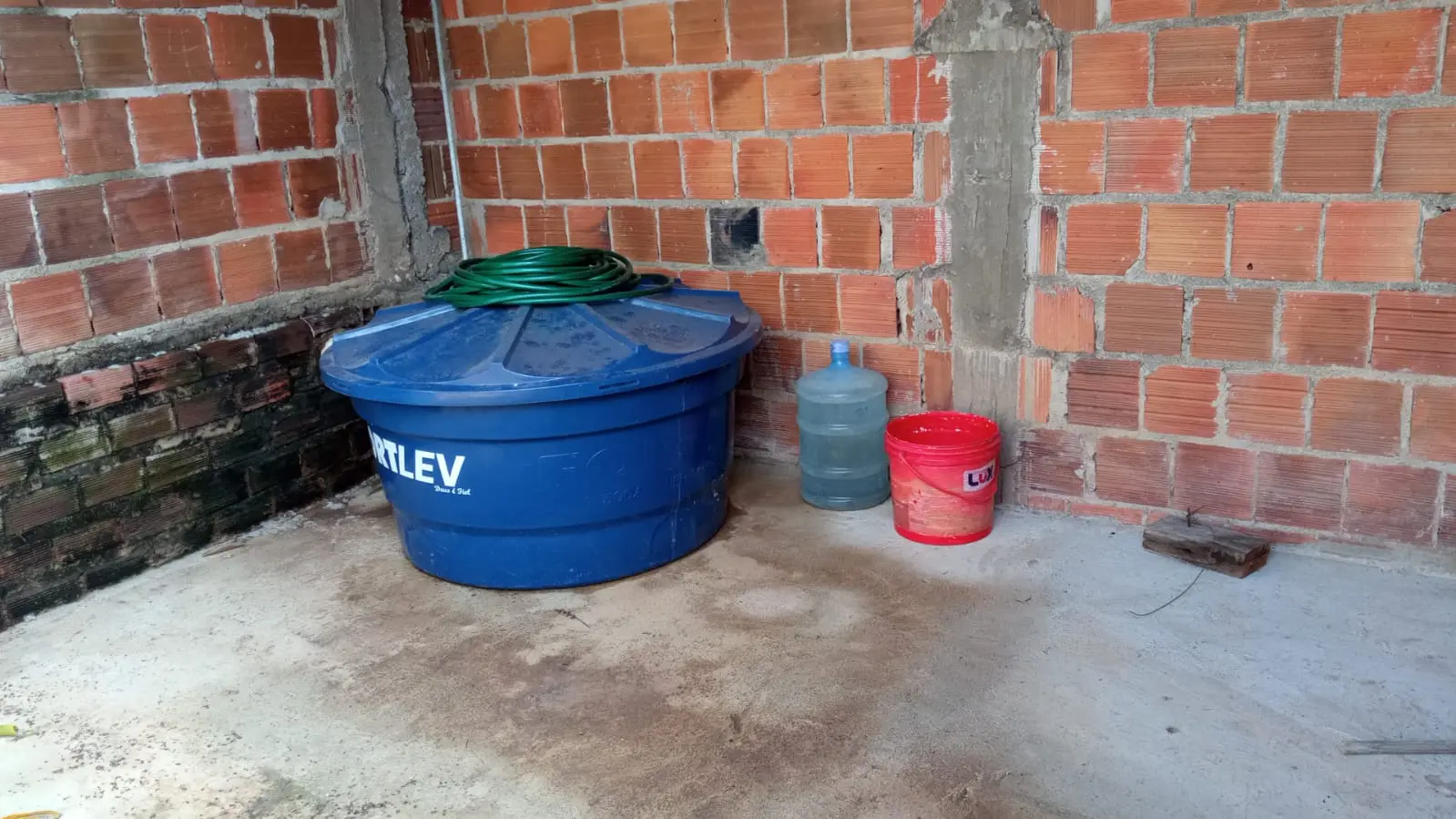 Imagem mostra o canto de um cômodo com paredes de tijolos expostos e colunas de concreto. No centro, há uma grande caixa dágua de plástico azul com uma mangueira enrolada em cima. Ao lado dela, à direita, estão um galão de água transparente e um balde vermelho. O chão é de cimento. Há um pequeno pedaço de madeira no chão, próximo à parede direita.