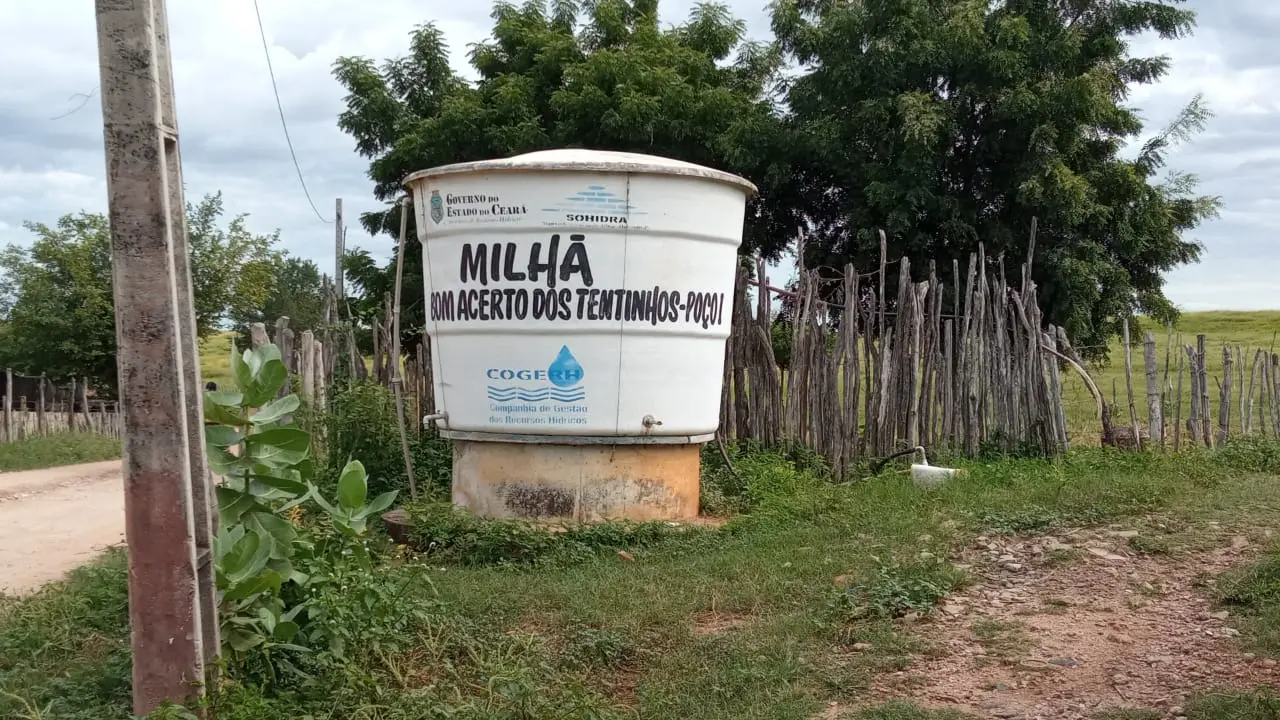 Imagem exibe um grande reservatório de água branco, cilíndrico. O reservatório está elevado sobre uma base de concreto e posicionado em uma área gramada. À direita, uma cerca rústica de madeira delimita um espaço, e atrás, árvores dominam a paisagem. Um poste de concreto se ergue no lado esquerdo da imagem, e um caminho de terra batida é visível no canto inferior esquerdo. O céu está claro com algumas nuvens.