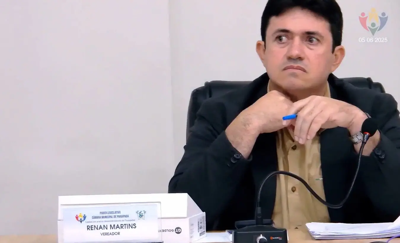 Imagem mostra o vereador Renan Martins, de Paraipaba, em sessão na Câmara Municipal, em meio a protestos de populares pela sua prisão. Ele está sentado na sua bancada no plenário. Veste um terno preto e uma camisa em tom ocre. Seus cabelos são escuros e sua pele clara.