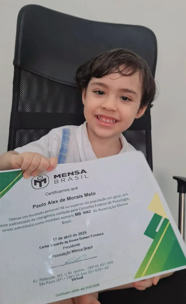 A imagem mostra um jovem menino de pele clara, com cabelo escuro e ondulado, sentado em uma cadeira de escritório preta. Ele está sorrindo diretamente para a câmera e segura um certificado grande com ambas as mãos, exibindo-o. O papel indica que Paulo Alex de Morais Melo é membro da Associação Mensa Brasil desde 17 de abril de 2025