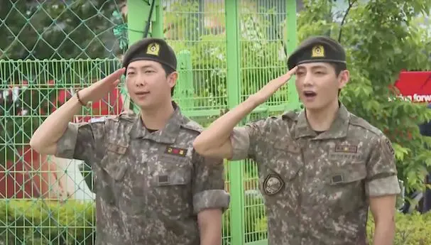 RM e V lado a lado, batendo continência. eles foram recebidos com flores e muito carinho dos fãs ao deixarem a base militar