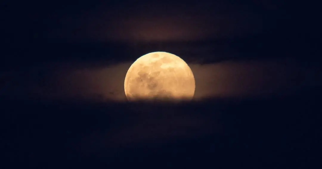 Imagem mostra uma grande Lua cheia de morango dourada se destaca mediante bancos de nuvens e névoa no céu noturno