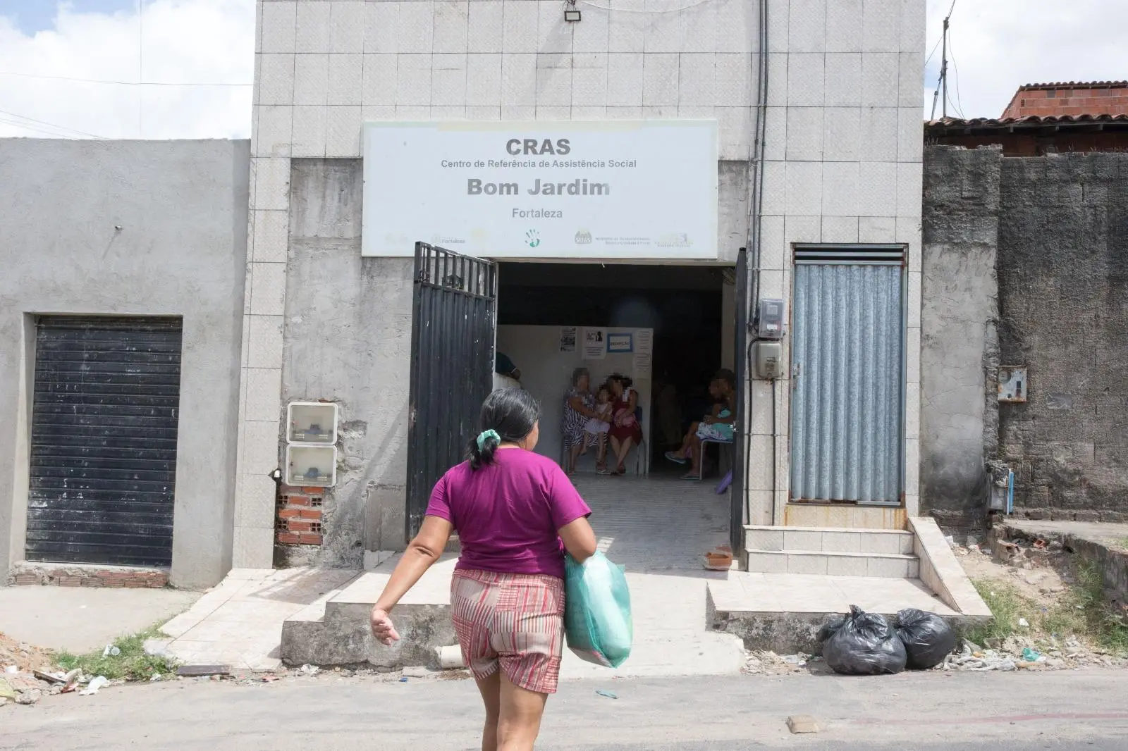 Imagem mostra fachada de um Centro de Referência em Assistência Psicossocial (CRAS) de Fortaleza. De costa para a imagem, em direção à porta, uma mulher com blusa rosa e short estampado em rosa e cinza.