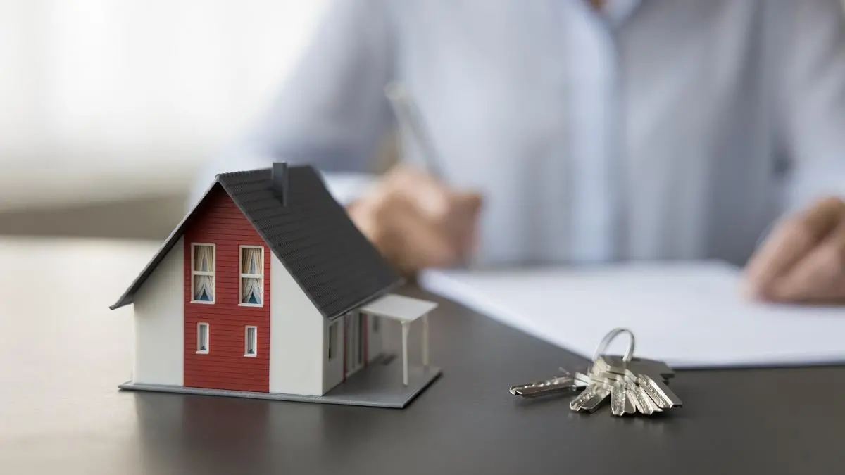 Casa em miniatura representando imóveis junto com uma chave e um homem assinando papel no fundo