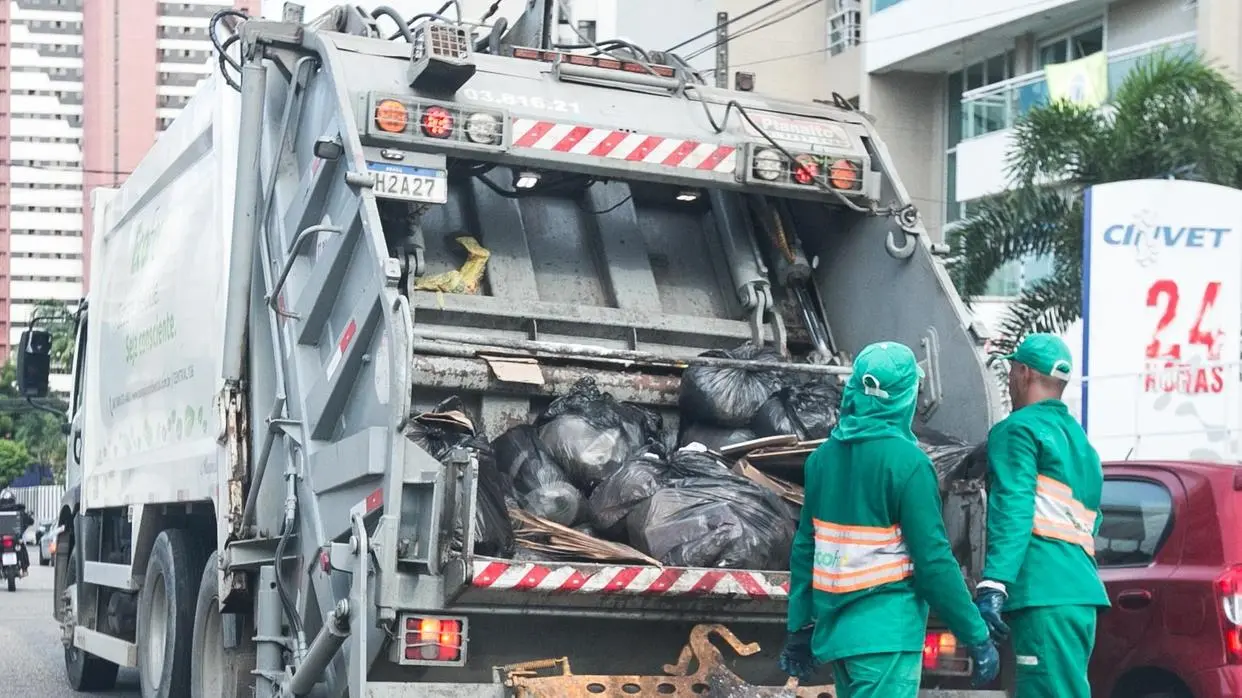 Foto mostra garis recolhendo lixo em uma rua de Fortaleza, com sacos pretos grandes em primeiro plano e um caminhão de coleta ao fundo, indicando uma ação de limpeza urbana em área residencial