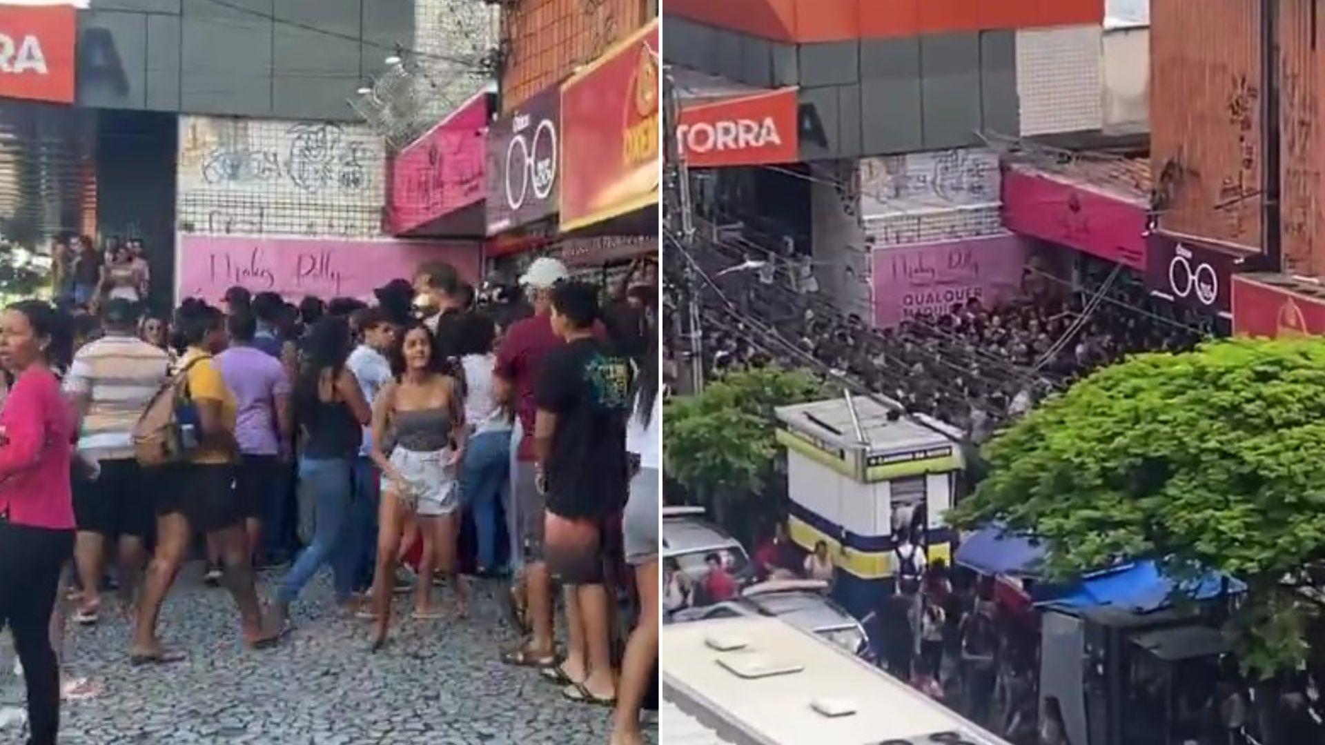 Fila grande de pessoas esperando em frente a uma loja no centro da cidade, com ruas movimentadas e lojas ao redor, durante o dia.