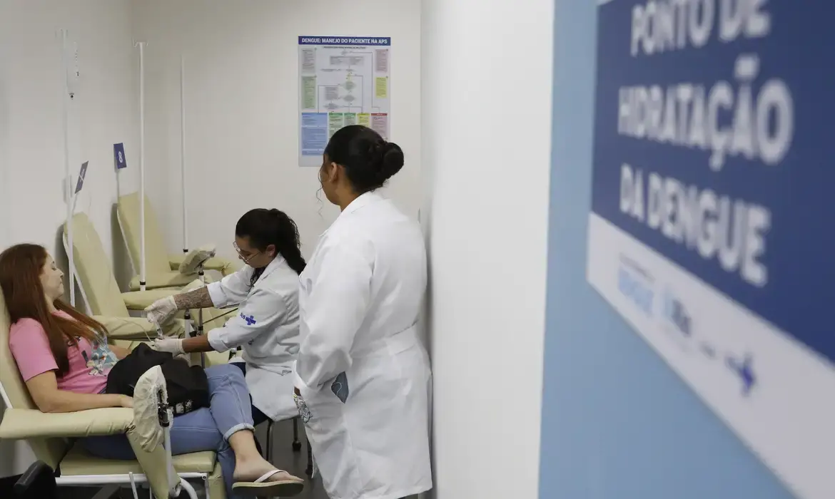 A imagem mostra duas profissionais de saúde atendendo uma paciente em uma unidade de saúde pública.