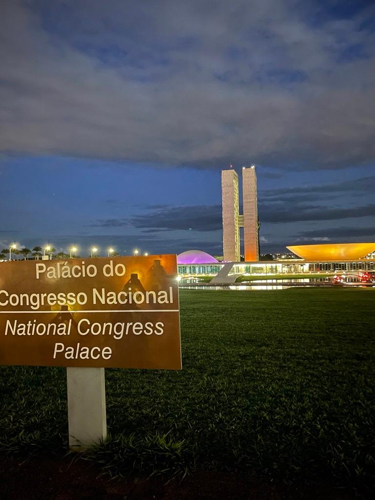 Foto do prédio do Congresso Nacional à noite, iluminado em laranja e roxo, com placa em português e inglês