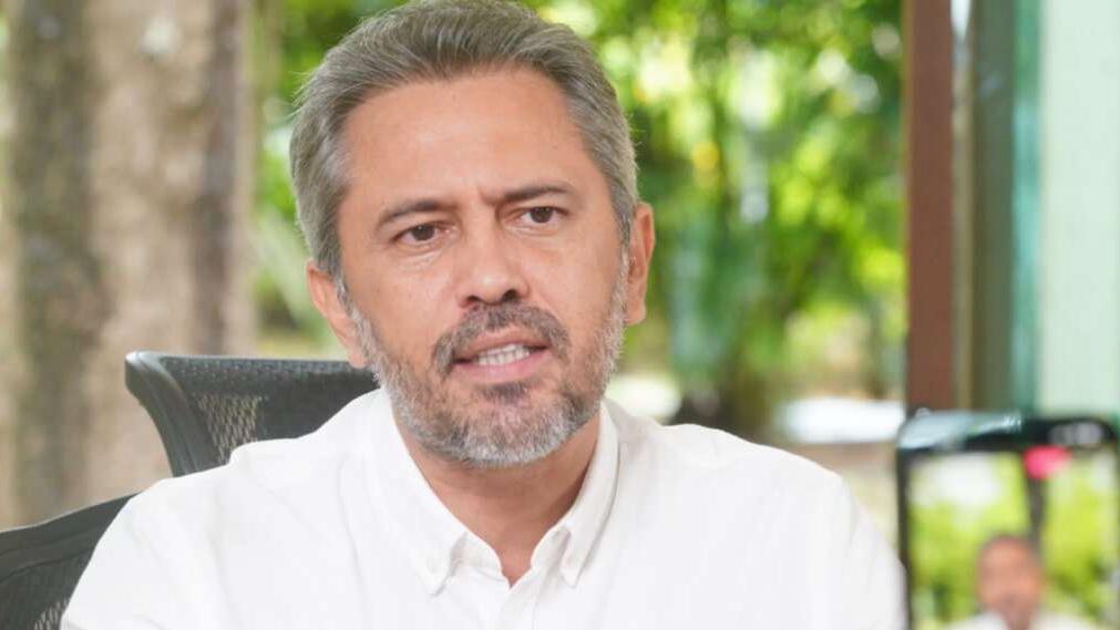 Elmano de Freitas, governador do Ceará, usando camisa branca, sentado em escritório com fundo de árvores verdes, gravando vídeo para redes sociais.