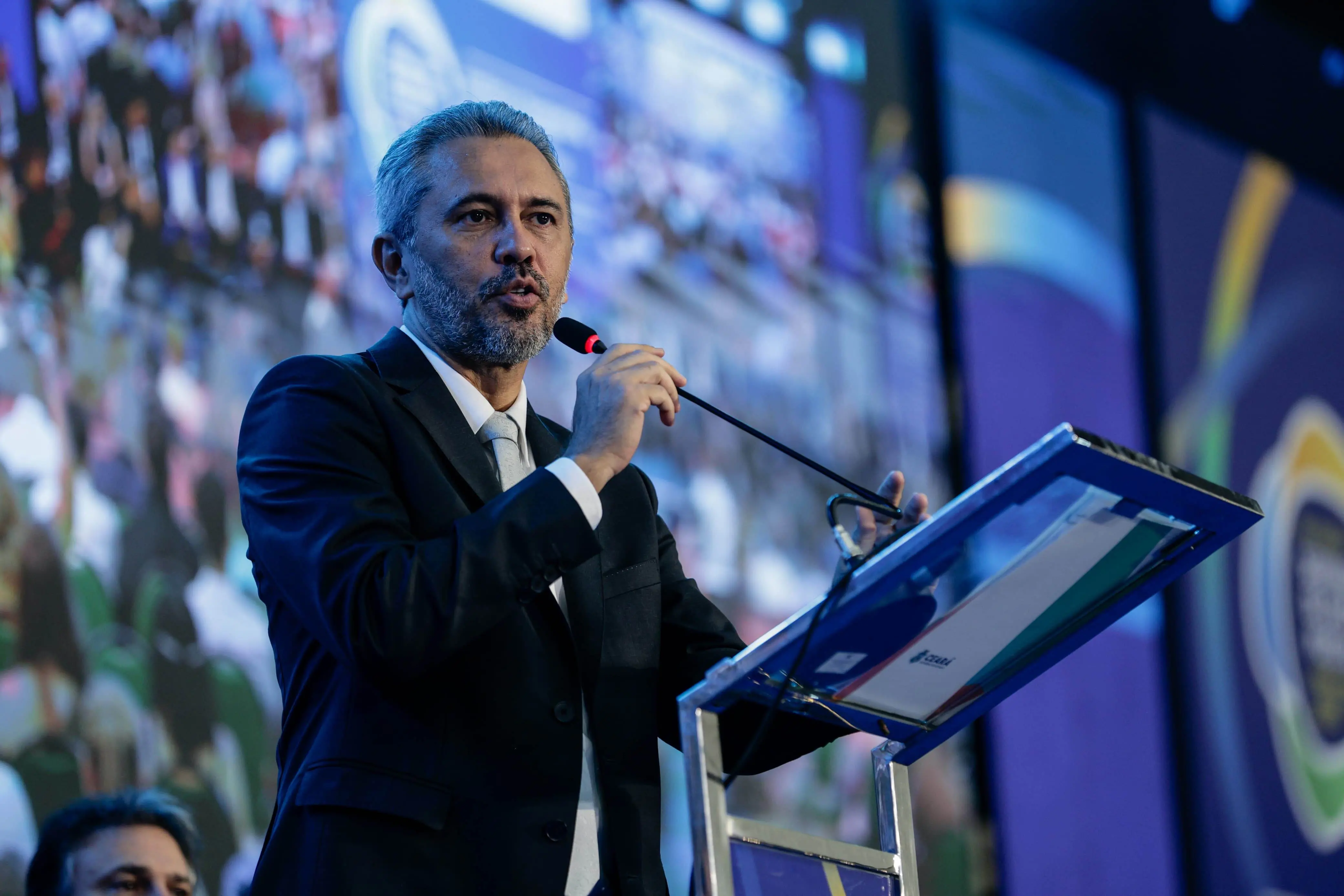 A imagem mostra o governador Elmano de Freitas falando ao microfone durante um painel no Seminário dos Prefeitos. Ele tem cabelos grisalhos e veste um terno escuro, com camisa branca e gravata cinza.