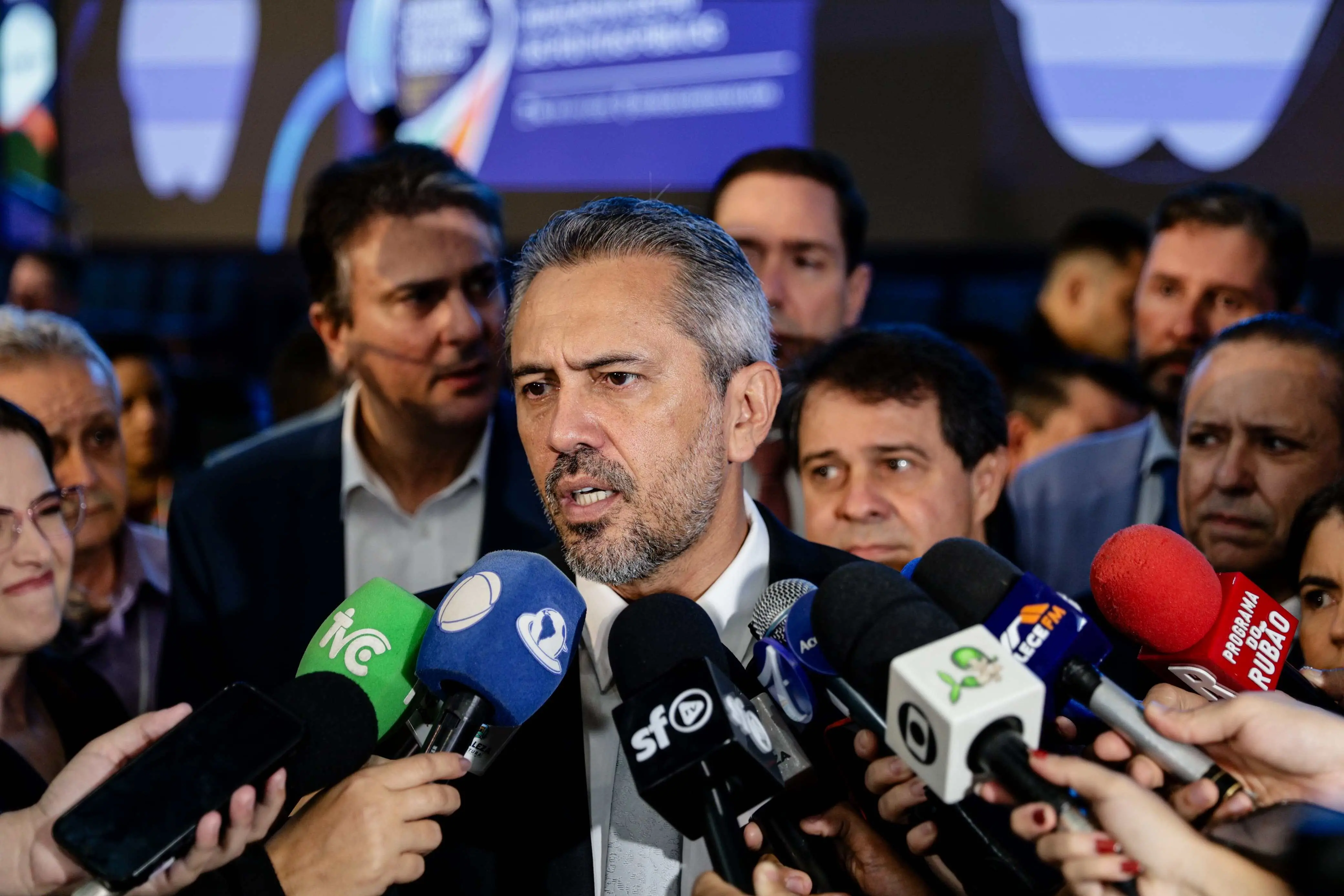 A imagem mostra o governador Elmano de Freitas durante entrevista coletiva na entrada do evento Seminário dos Prefeitos, no Ceará. Ela fala a jornalistas. Elmano veste um terno escuro, camisa branca e gravata cinza. Tem cabelo grisalho.