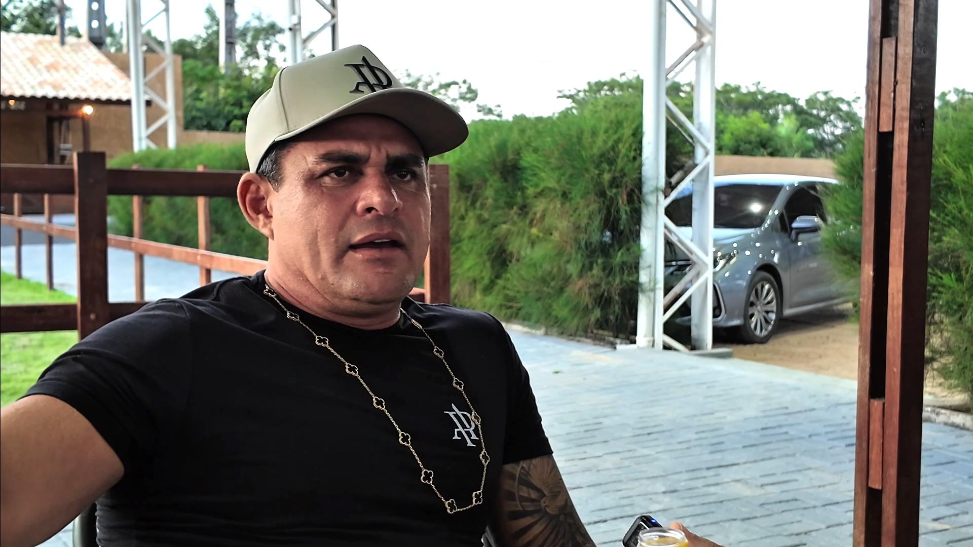 Isaías CDs em casa durante entrevista ao comunicador Gordo Show. Ele usa blusa preta e uma colar, além deum boné